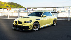 Pastel yellow BMW G82 M4 with carbon fiber front splitter, hood vents, side canards, bronze wheels, and large rear wing parked at a race track facility during golden hour.