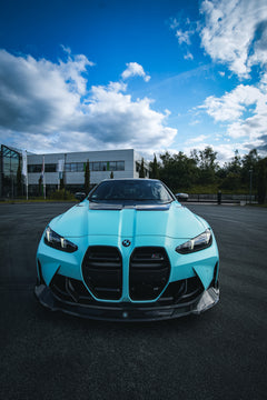 Turquoise BMW with carbon fiber body kit parked outdoors, highlighting aggressive front lip, vented hood, and dramatic sky backdrop.