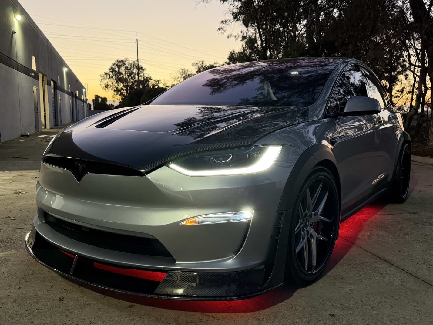 Tesla Model X featuring an aggressive REVOZPORT carbon fiber body kit with front lip, vented hood, wide fender extensions, side skirts, and illuminated red underglow lighting at dusk.