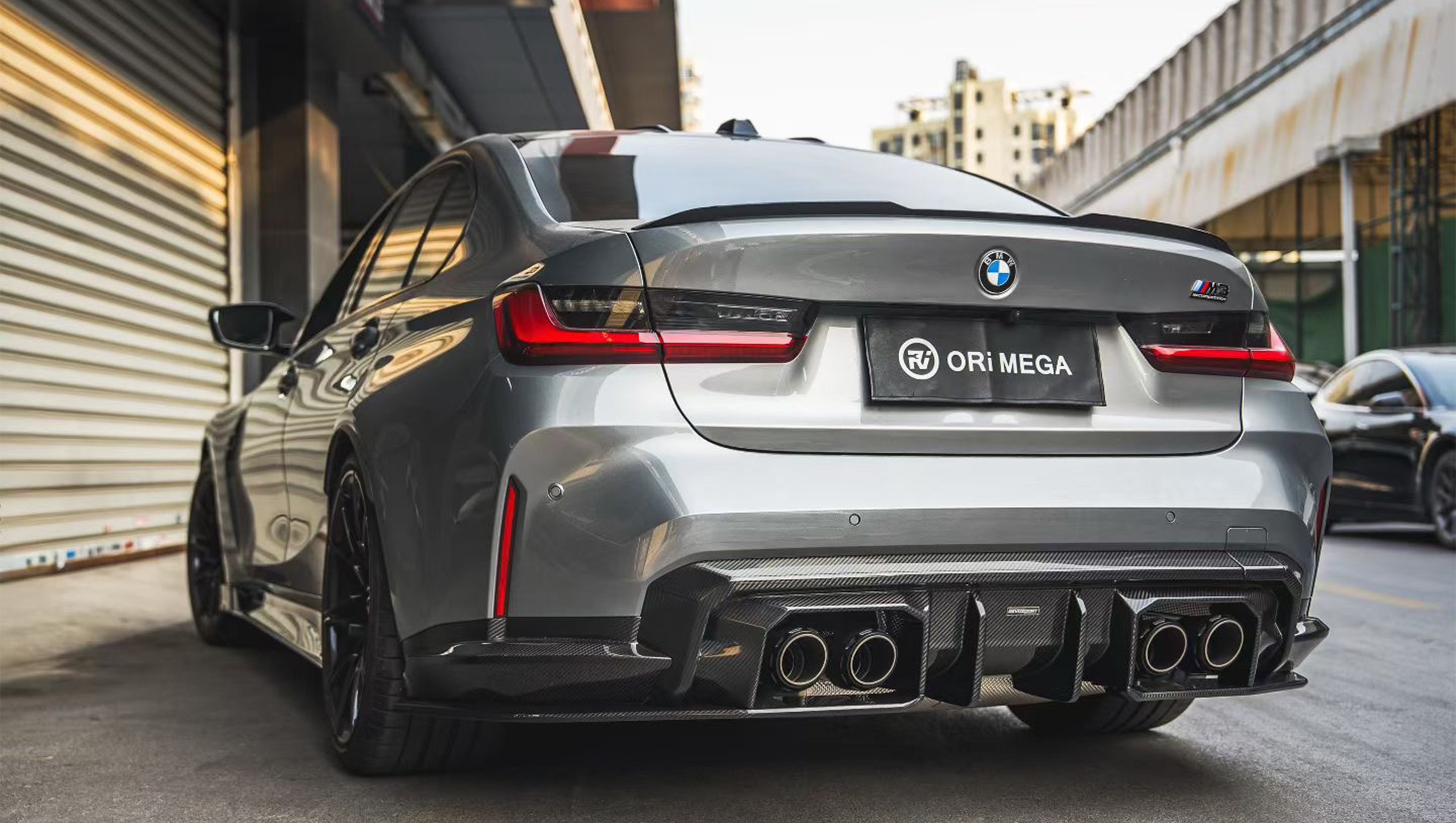 Gray BMW G80 M3 featuring a REVOZPORT carbon fiber front lip, side skirts, and aggressive aero styling parked outside a modern urban garage.