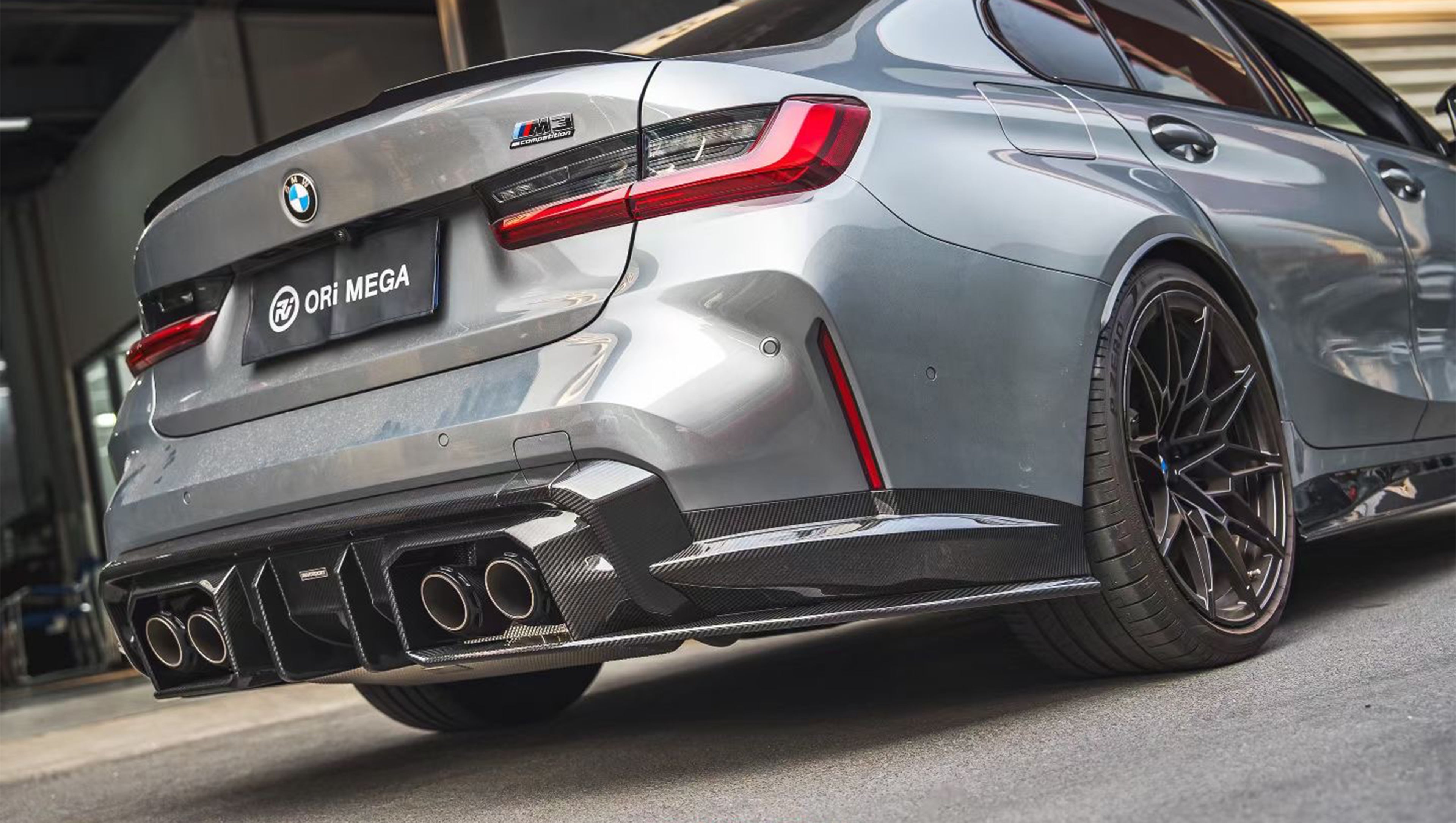Gray BMW G80 M3 featuring a REVOZPORT carbon fiber front lip, side skirts, and aggressive aero styling parked outside a modern urban garage.