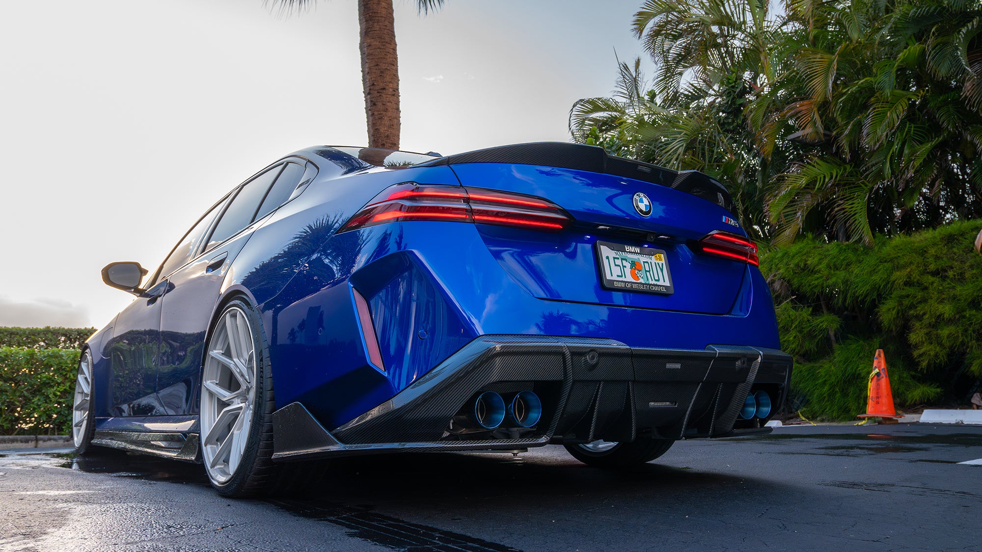 Blue BMW G90 M5 featuring a REVOZPORT carbon fiber front lip, side skirts, aggressive aero styling, and silver forged wheels parked on a wet road with palm trees at sunset.