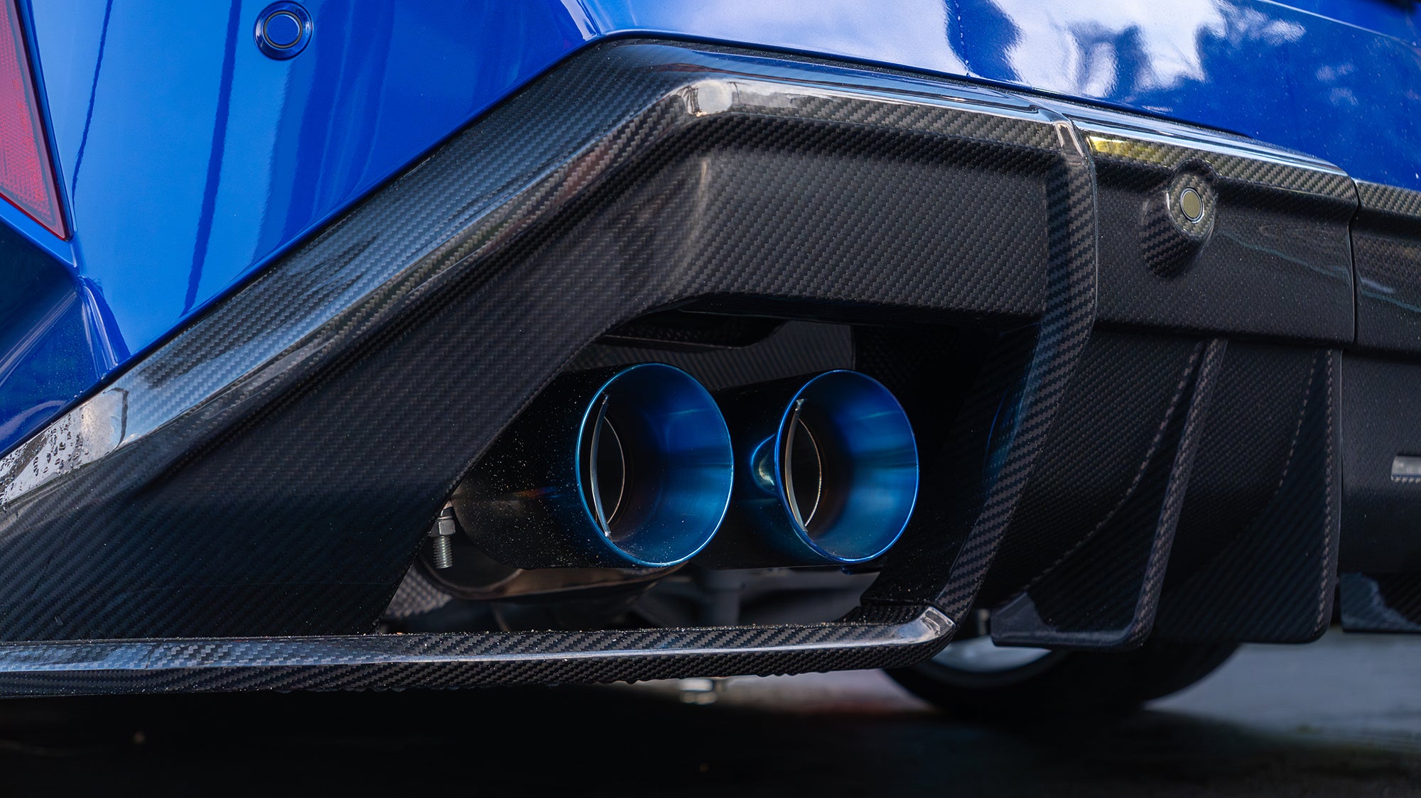 Blue BMW G90 M5 featuring a REVOZPORT carbon fiber front lip, side skirts, aggressive aero styling, and silver forged wheels parked on a wet road with palm trees at sunset.