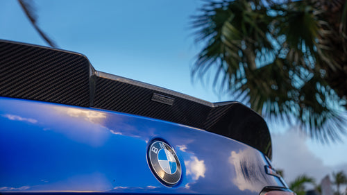 Blue BMW G90 M5 featuring a REVOZPORT carbon fiber front lip, side skirts, aggressive aero styling, and silver forged wheels parked on a wet road with palm trees at sunset.