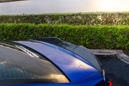 Blue BMW G90 M5 featuring a REVOZPORT carbon fiber front lip, side skirts, aggressive aero styling, and silver forged wheels parked on a wet road with palm trees at sunset.