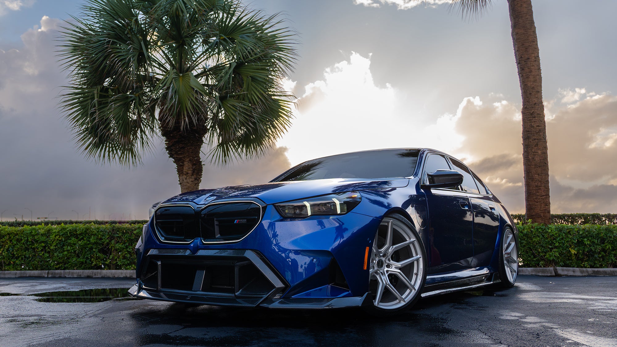 Blue BMW G90 M5 featuring a REVOZPORT carbon fiber front lip, side skirts, aggressive aero styling, and silver forged wheels parked on a wet road with palm trees at sunset.