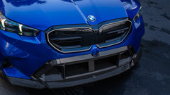 Blue BMW G90 M5 featuring a REVOZPORT carbon fiber front lip, side skirts, aggressive aero styling, and silver forged wheels parked on a wet road with palm trees at sunset.