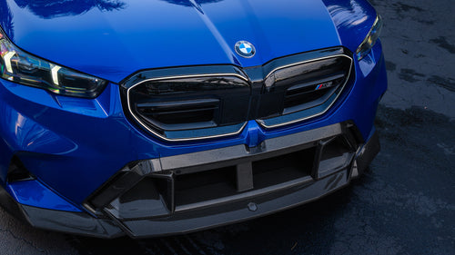 Blue BMW G90 M5 featuring a REVOZPORT carbon fiber front lip, side skirts, aggressive aero styling, and silver forged wheels parked on a wet road with palm trees at sunset.
