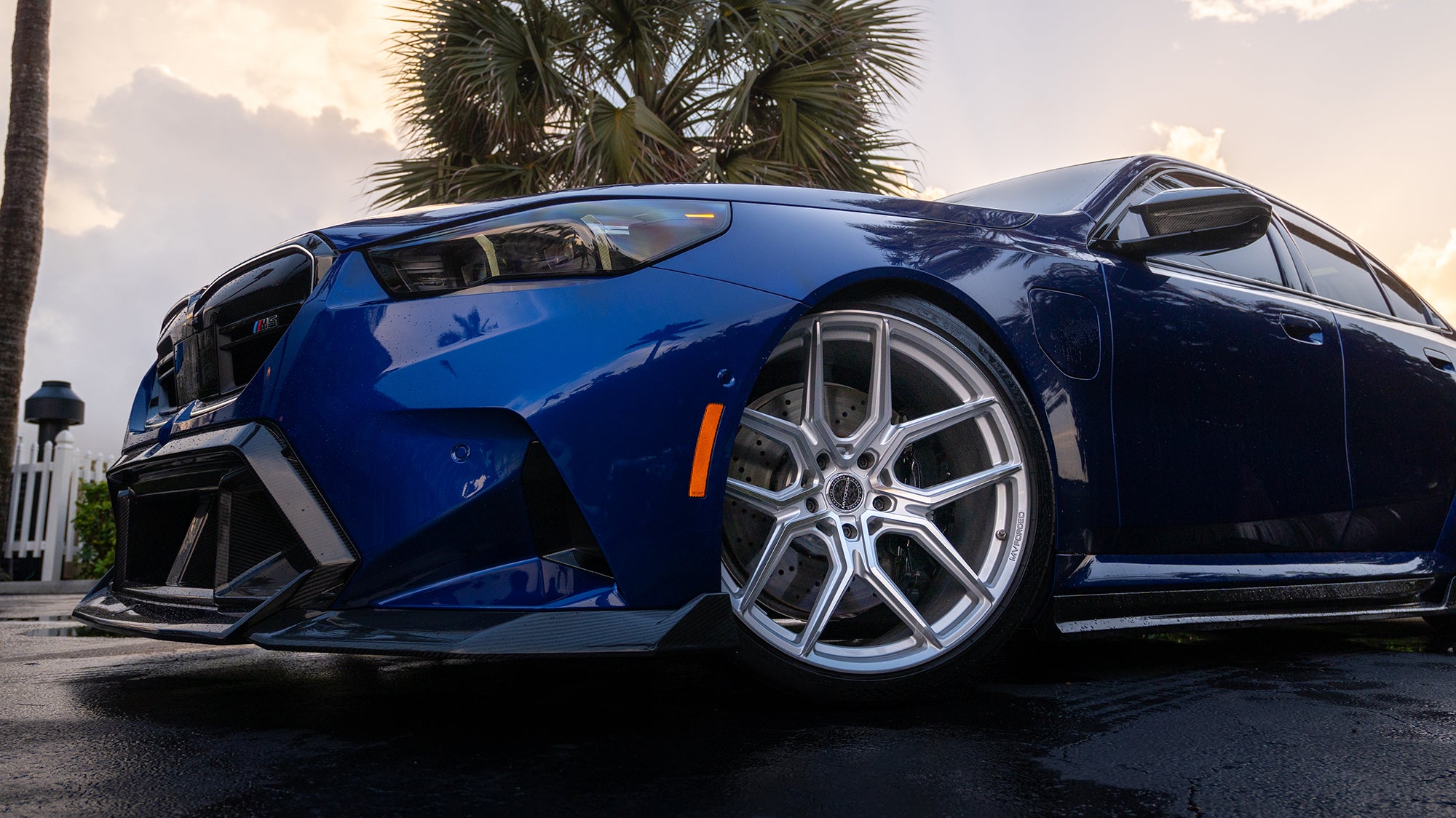 Blue BMW G90 M5 featuring a REVOZPORT carbon fiber front lip, side skirts, aggressive aero styling, and silver forged wheels parked on a wet road with palm trees at sunset.