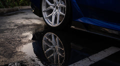 Blue BMW G90 M5 featuring a REVOZPORT carbon fiber front lip, side skirts, aggressive aero styling, and silver forged wheels parked on a wet road with palm trees at sunset.