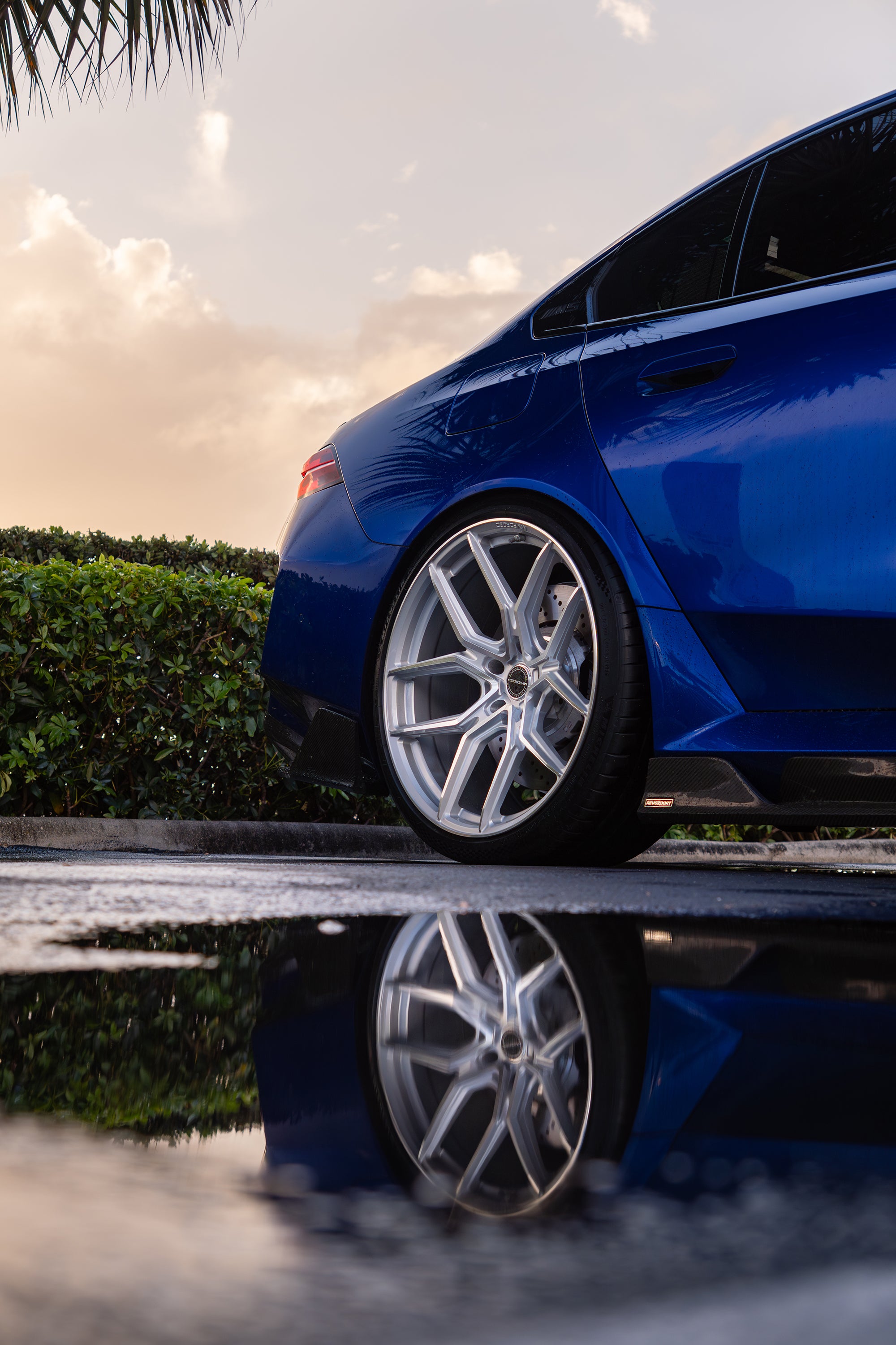 Blue BMW G90 M5 featuring a REVOZPORT carbon fiber front lip, side skirts, aggressive aero styling, and silver forged wheels parked on a wet road with palm trees at sunset.