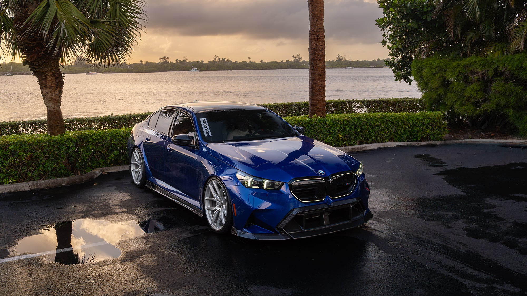 Blue BMW G90 M5 featuring a REVOZPORT carbon fiber front lip, side skirts, aggressive aero styling, and silver forged wheels parked on a wet road with palm trees at sunset.