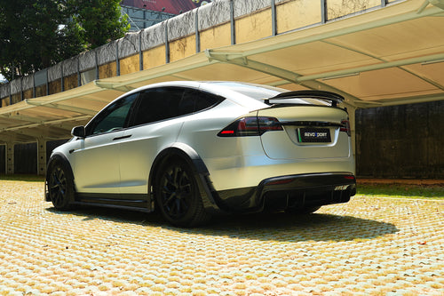 Silver Tesla Model X featuring a REVOZPORT body kit with carbon fiber front lip, side skirts, wide fender flares, and lowered stance parked outdoors in sunlight.