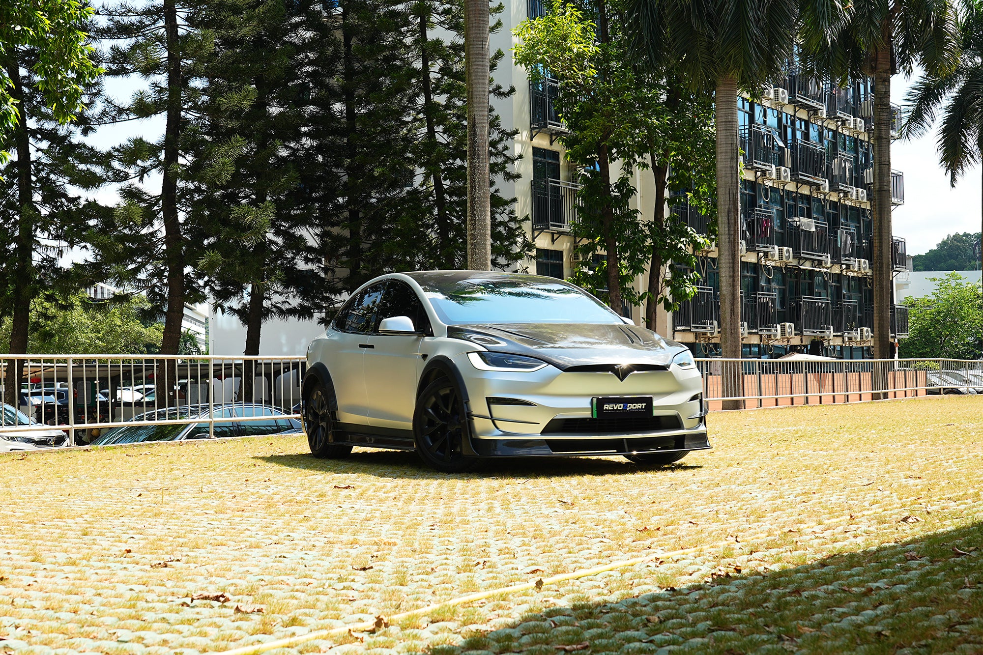 Silver Tesla Model X featuring a REVOZPORT body kit with carbon fiber front lip, side skirts, wide fender flares, and lowered stance parked outdoors in sunlight.