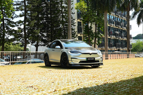 Silver Tesla Model X featuring a REVOZPORT body kit with carbon fiber front lip, side skirts, wide fender flares, and lowered stance parked outdoors in sunlight.