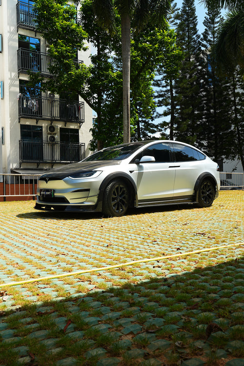 Silver Tesla Model X featuring a REVOZPORT body kit with carbon fiber front lip, side skirts, wide fender flares, and lowered stance parked outdoors in sunlight.
