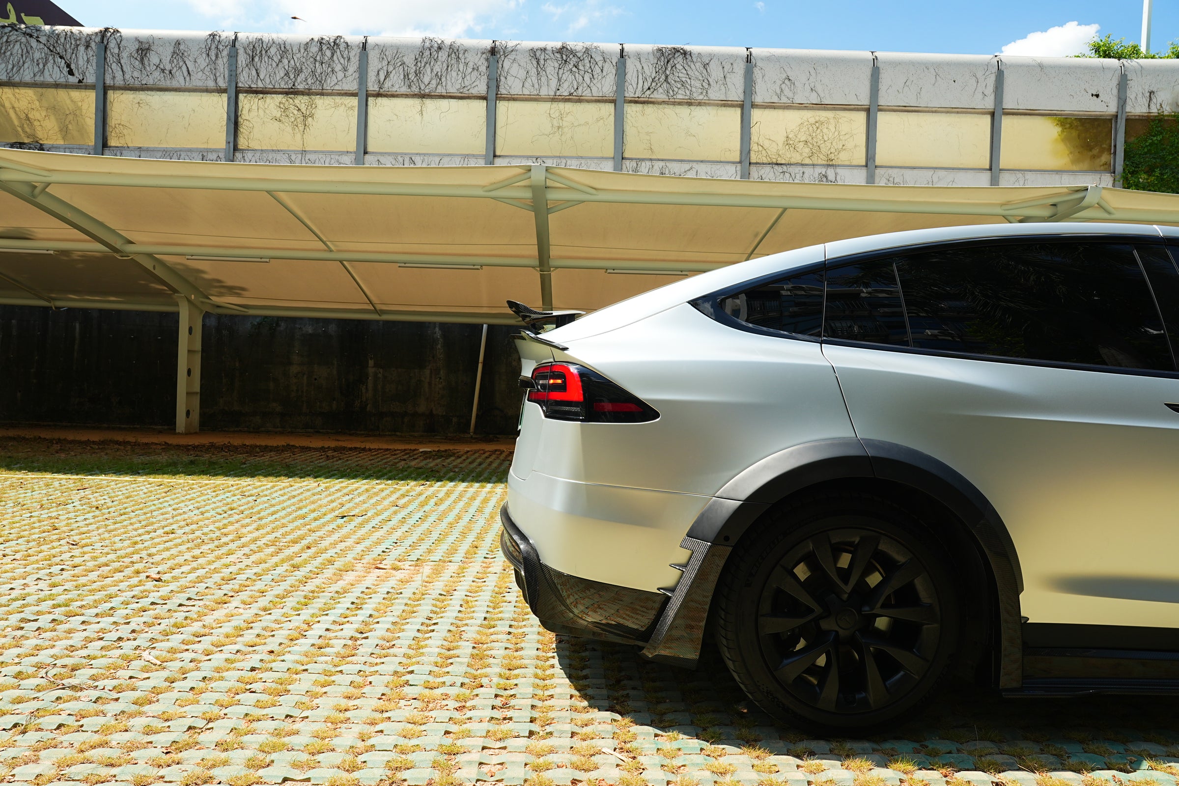 Silver Tesla Model X featuring a REVOZPORT body kit with carbon fiber front lip, side skirts, wide fender flares, and lowered stance parked outdoors in sunlight.