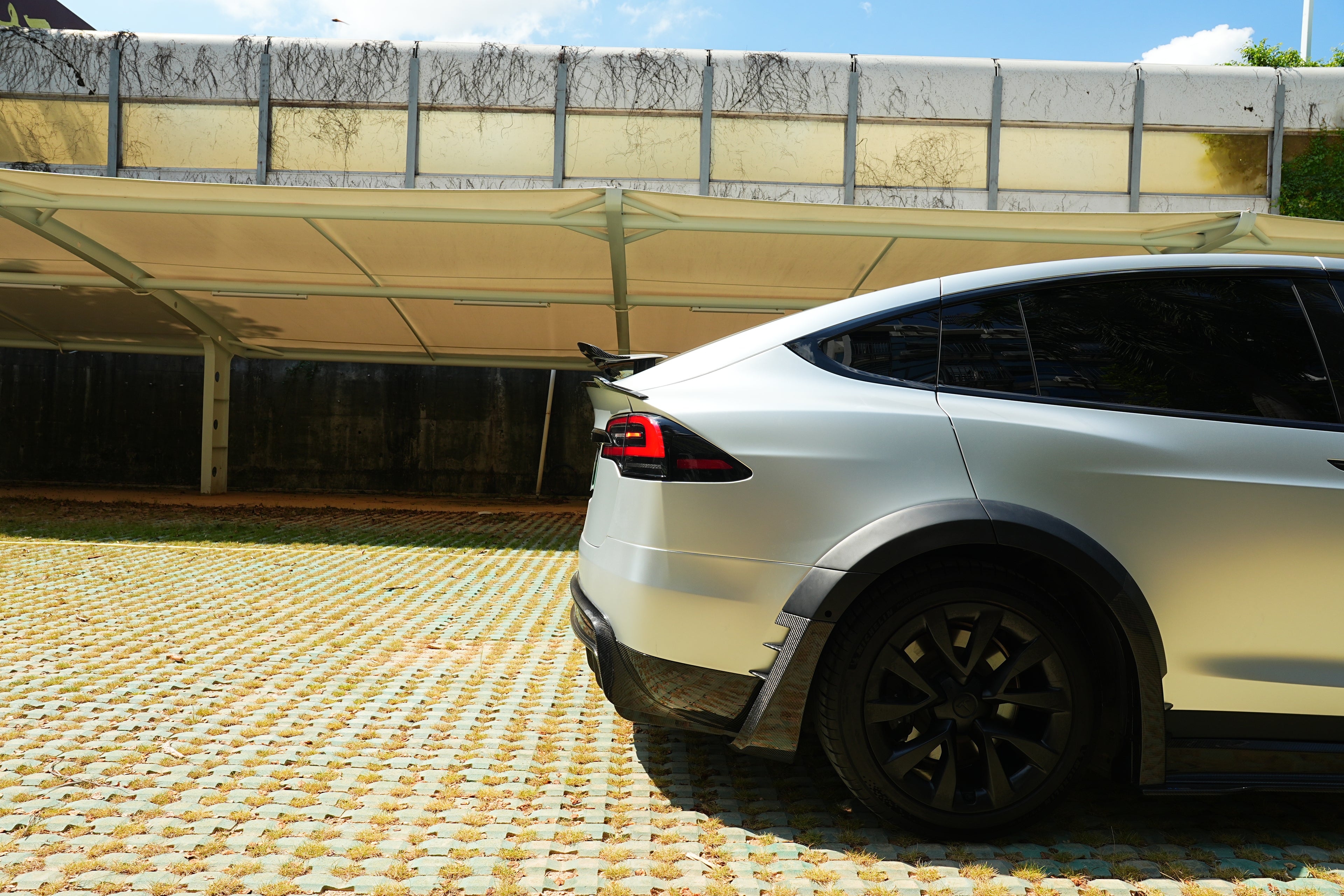 Silver Tesla Model X featuring a REVOZPORT body kit with carbon fiber front lip, side skirts, wide fender flares, and lowered stance parked outdoors in sunlight.