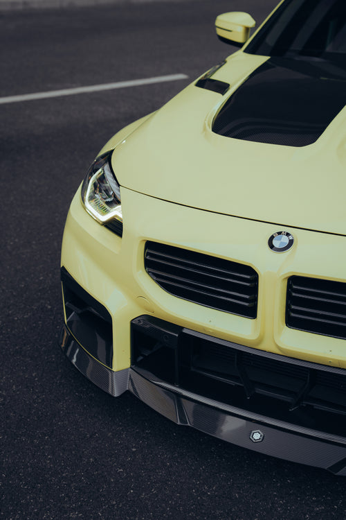 Close-up of a yellow BMW with carbon fiber front lip and hood vents, highlighting aggressive front aero styling.