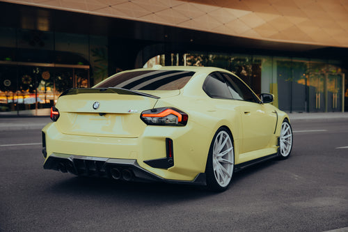 Yellow BMW with carbon fiber rear spoiler, diffuser, and side skirts parked outside a modern building, emphasizing rear aero styling.