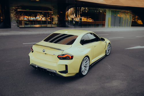 Yellow BMW with carbon fiber rear spoiler, diffuser, and side skirts parked outside a modern building, emphasizing sleek rear styling and stance.