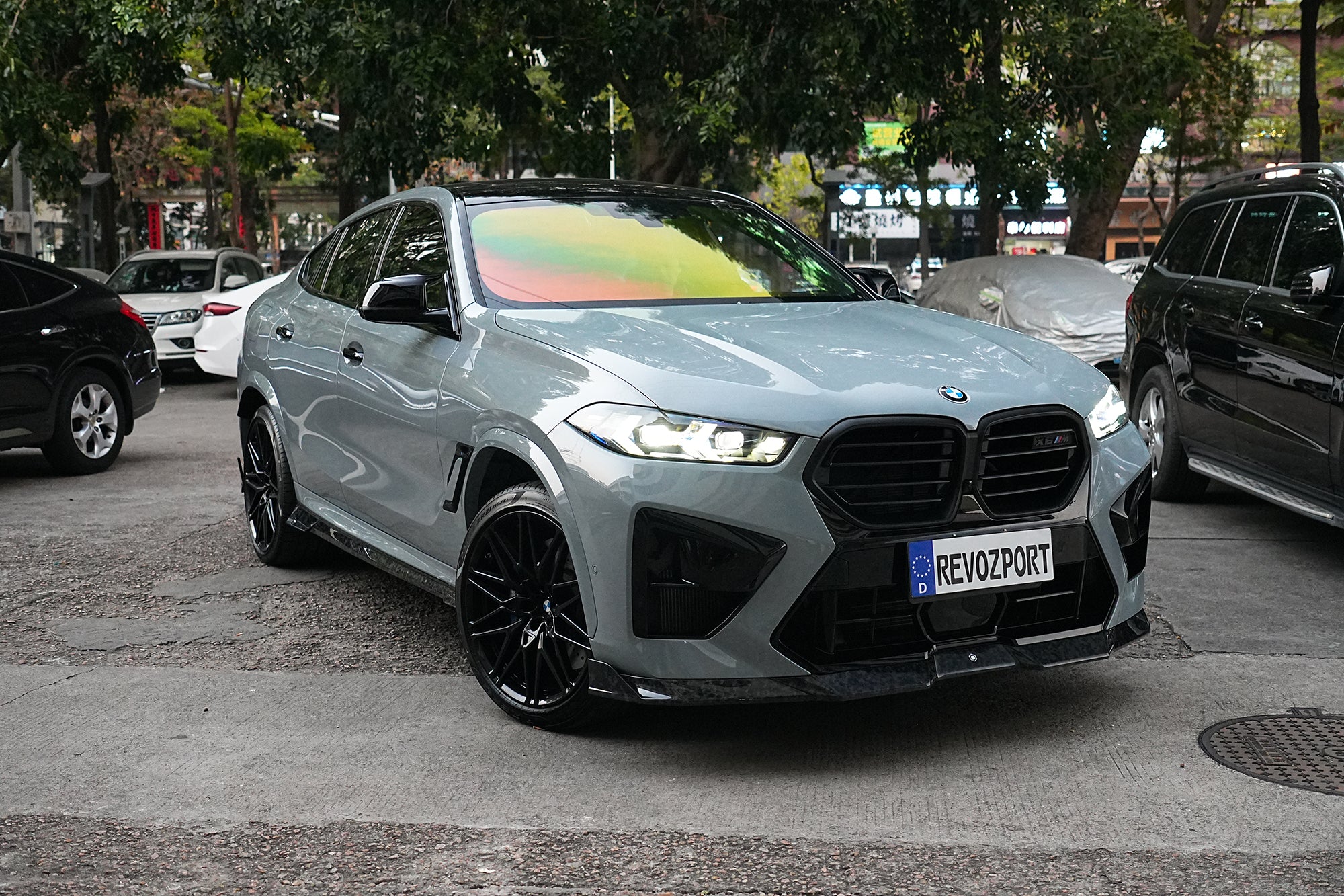Gray BMW X6M featuring a REVOZPORT carbon fiber body kit with aggressive front lip, side skirts, and sporty aero styling parked on an urban street at dusk.