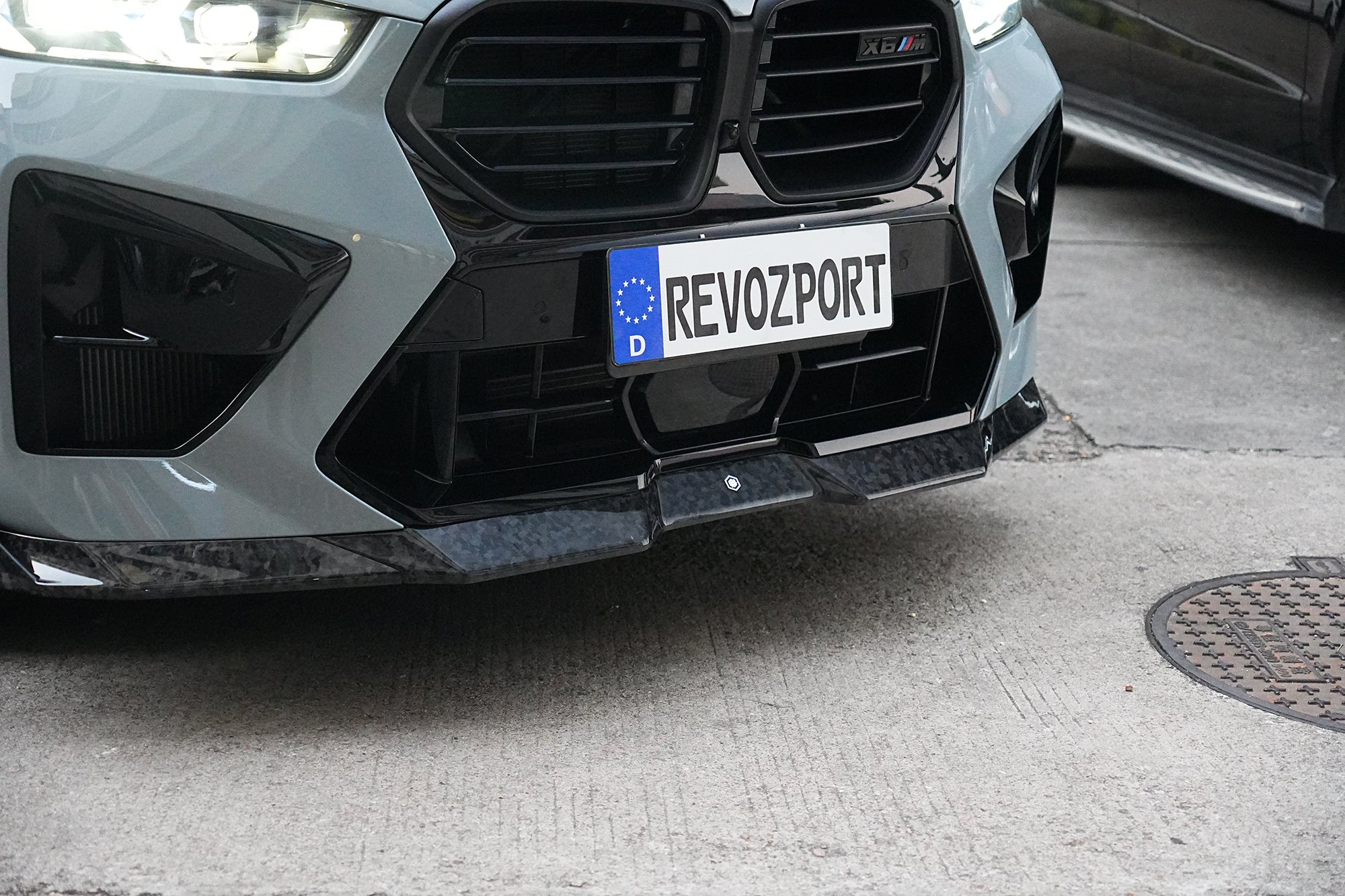 Gray BMW X6M featuring a REVOZPORT carbon fiber body kit with aggressive front lip, side skirts, and sporty aero styling parked on an urban street at dusk.