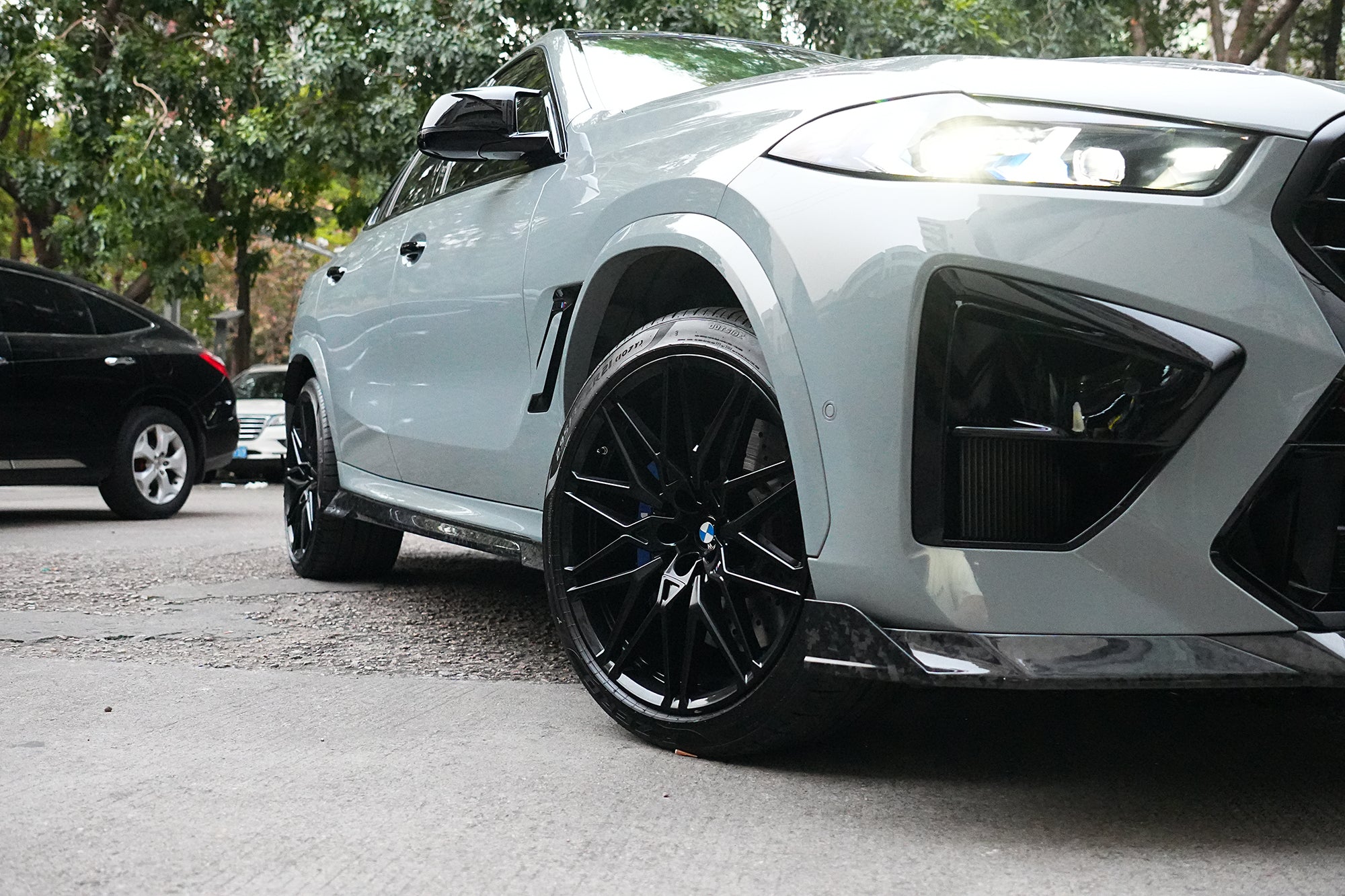 Gray BMW X6M featuring a REVOZPORT carbon fiber body kit with aggressive front lip, side skirts, and sporty aero styling parked on an urban street at dusk.