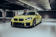 The BMW M2, wrapped in carbon fiber, sits in a high-end studio with lights on, equipped with high-end wheels, side skirts, and front lip