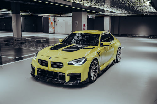 Yellow BMW with carbon fiber body kit parked in a modern garage, highlighting front lip, hood vents, and side skirt styling.