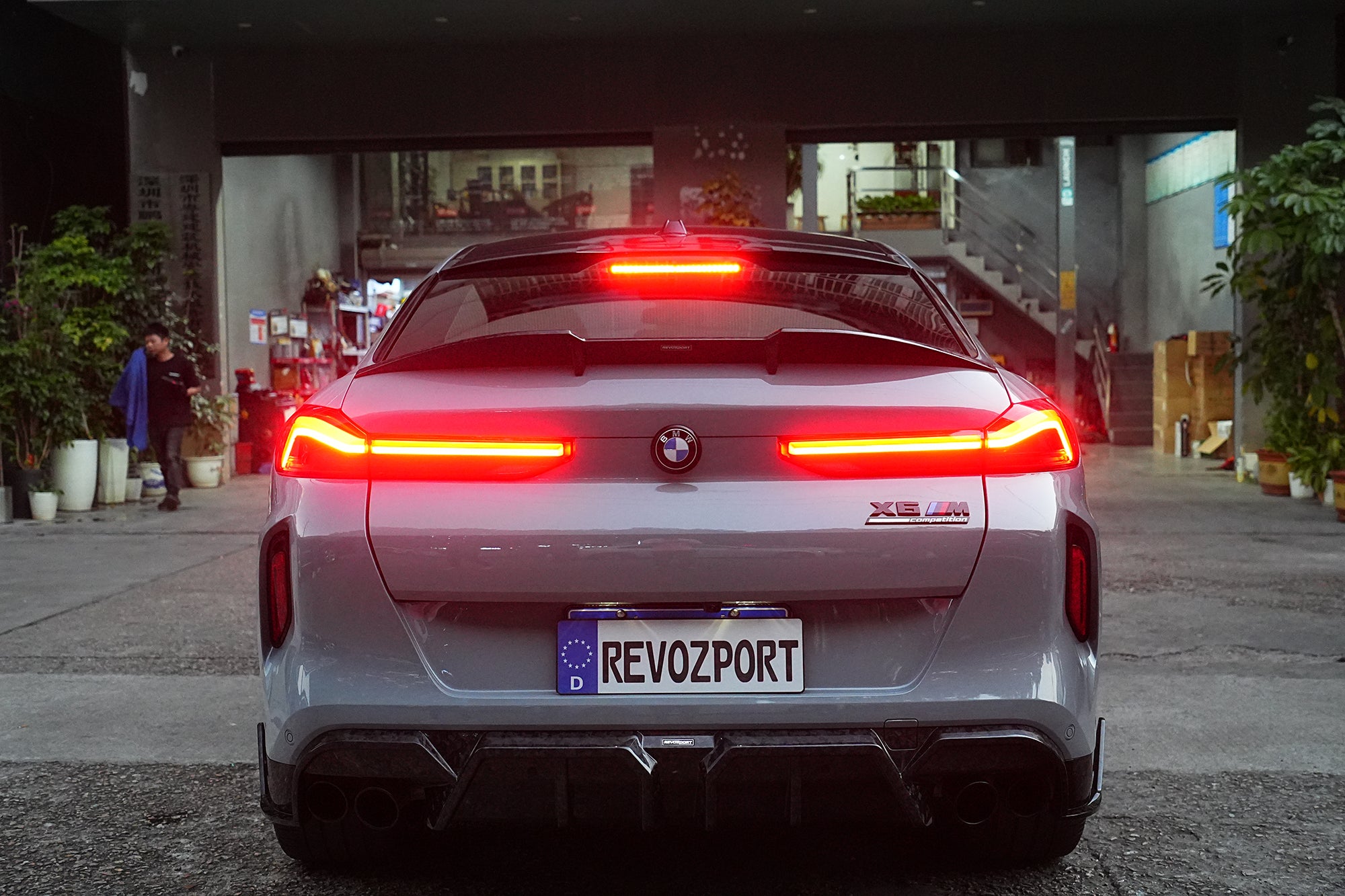 Gray BMW X6M featuring a REVOZPORT carbon fiber body kit with aggressive front lip, side skirts, and sporty aero styling parked on an urban street at dusk.