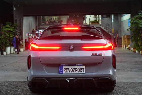 Gray BMW X6M featuring a REVOZPORT carbon fiber body kit with aggressive front lip, side skirts, and sporty aero styling parked on an urban street at dusk.
