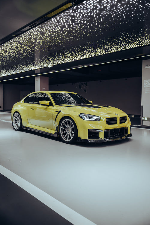 The BMW M2, wrapped in carbon fiber, sits in a high-end studio with lights on, equipped with high-end wheels, side skirts, and front lip