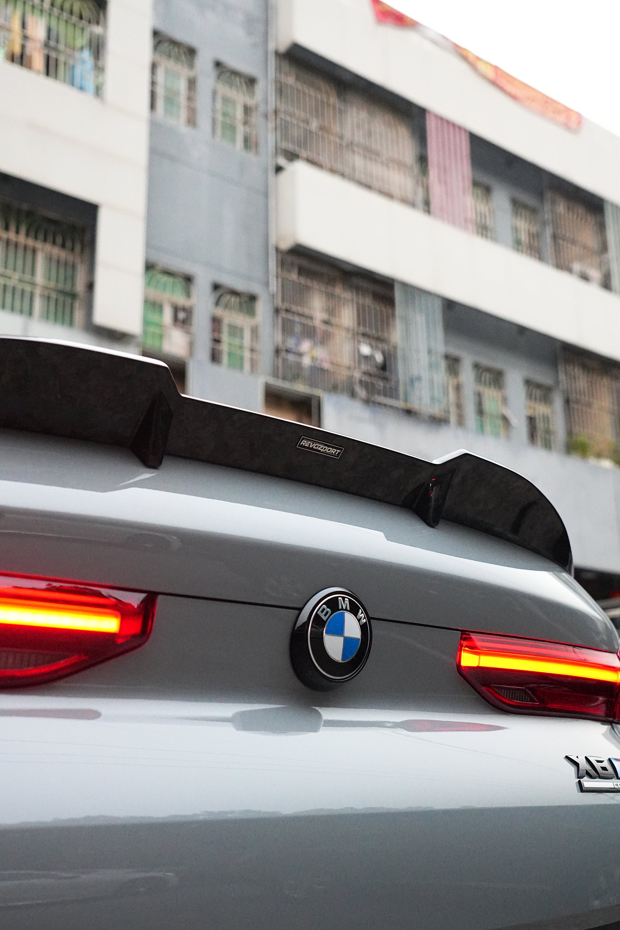 Gray BMW X6M featuring a REVOZPORT carbon fiber body kit with aggressive front lip, side skirts, and sporty aero styling parked on an urban street at dusk.