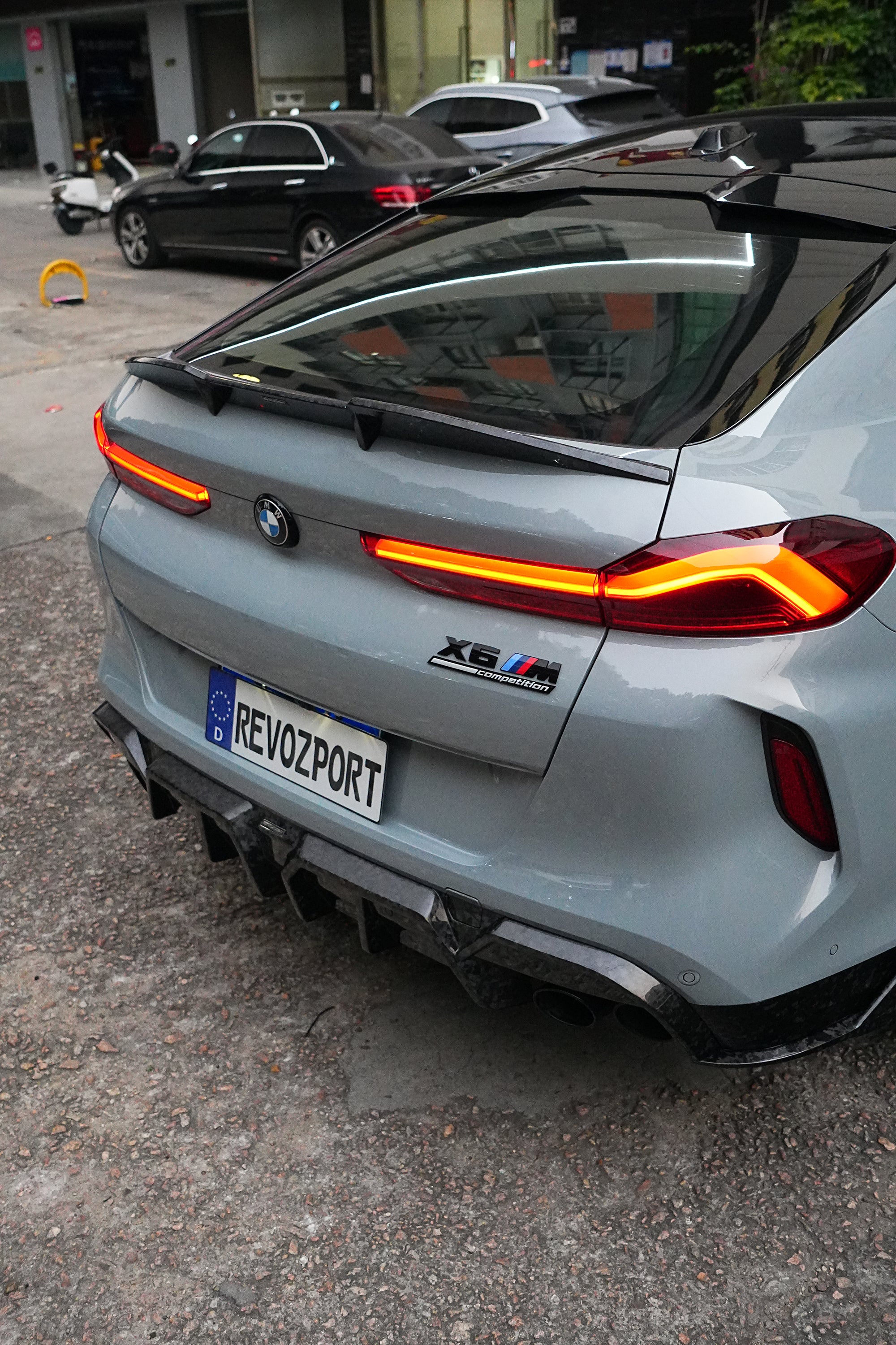 Gray BMW X6M featuring a REVOZPORT carbon fiber body kit with aggressive front lip, side skirts, and sporty aero styling parked on an urban street at dusk.