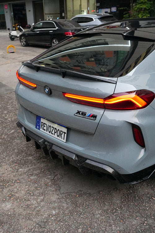 Gray BMW X6M featuring a REVOZPORT carbon fiber body kit with aggressive front lip, side skirts, and sporty aero styling parked on an urban street at dusk.