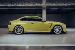 A close-up of the side of a BMW M2, wrapped in carbon fiber, in a high-end studio with lights on, equipped with a front grille, front lip, hood, wheel hubs, and side skirts,