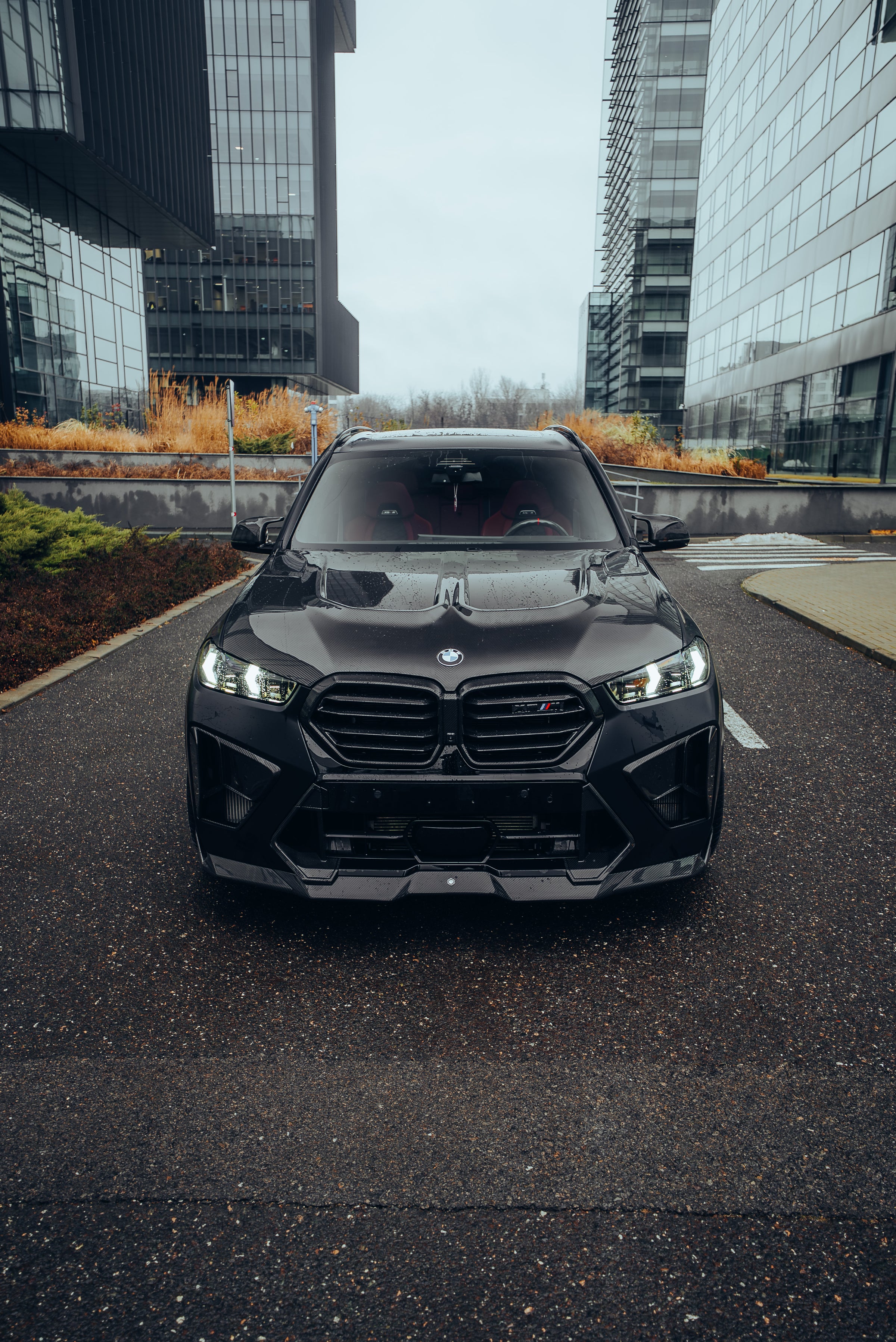 Black BMW X5M featuring a REVOZPORT carbon fiber body kit with aggressive front lip, side skirts, enlarged front air intakes, and sporty aero styling parked in a modern urban business district.