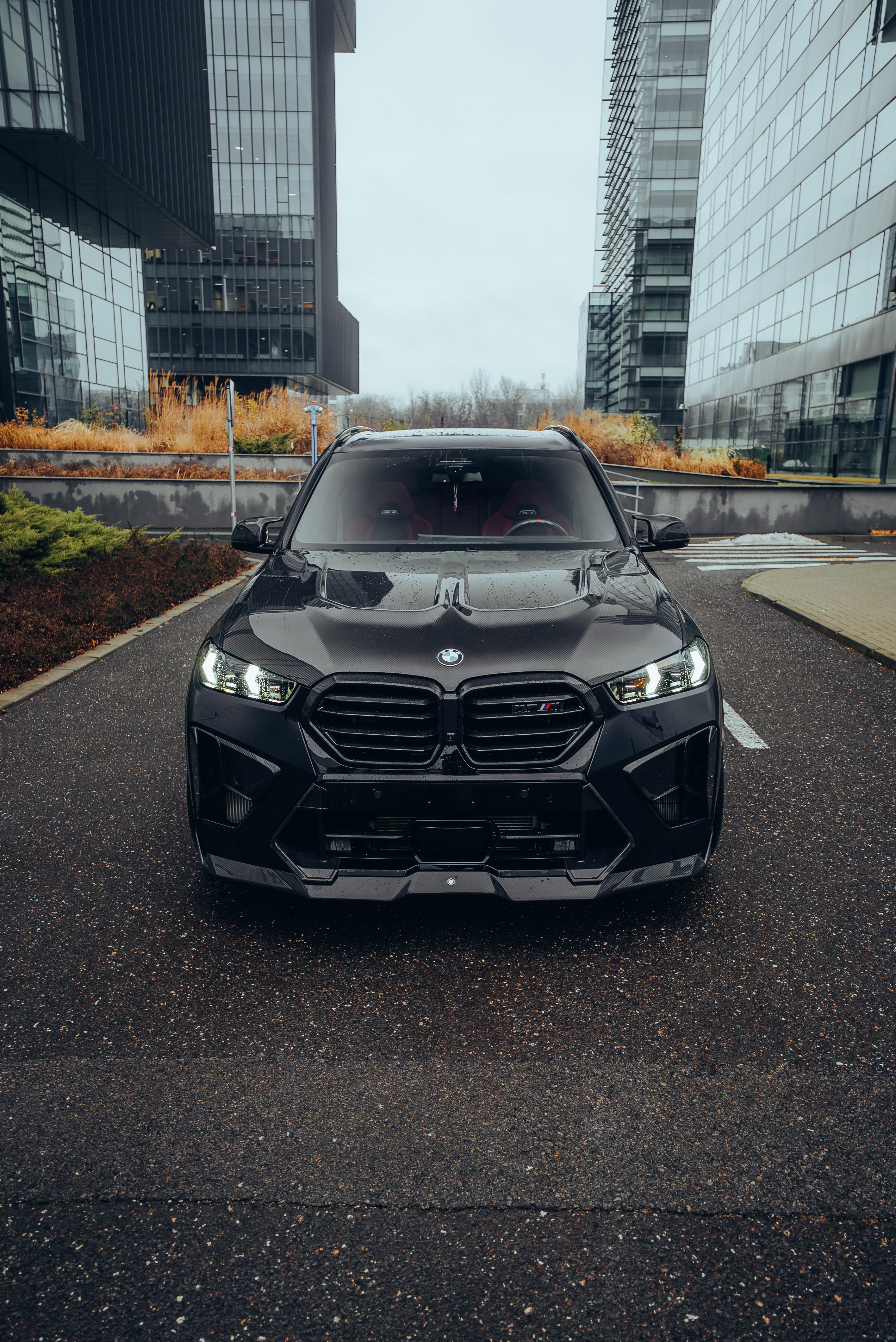 Black BMW X5M featuring a REVOZPORT carbon fiber body kit with aggressive front lip, side skirts, enlarged front air intakes, and sporty aero styling parked in a modern urban business district.