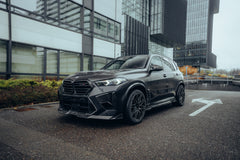 Black BMW X5M featuring a REVOZPORT carbon fiber body kit with aggressive front lip, side skirts, enlarged front air intakes, and sporty aero styling parked in a modern urban business district.