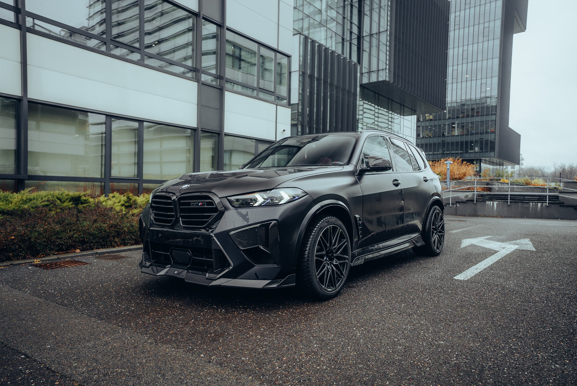 Black BMW X5M featuring a REVOZPORT carbon fiber body kit with aggressive front lip, side skirts, enlarged front air intakes, and sporty aero styling parked in a modern urban business district.