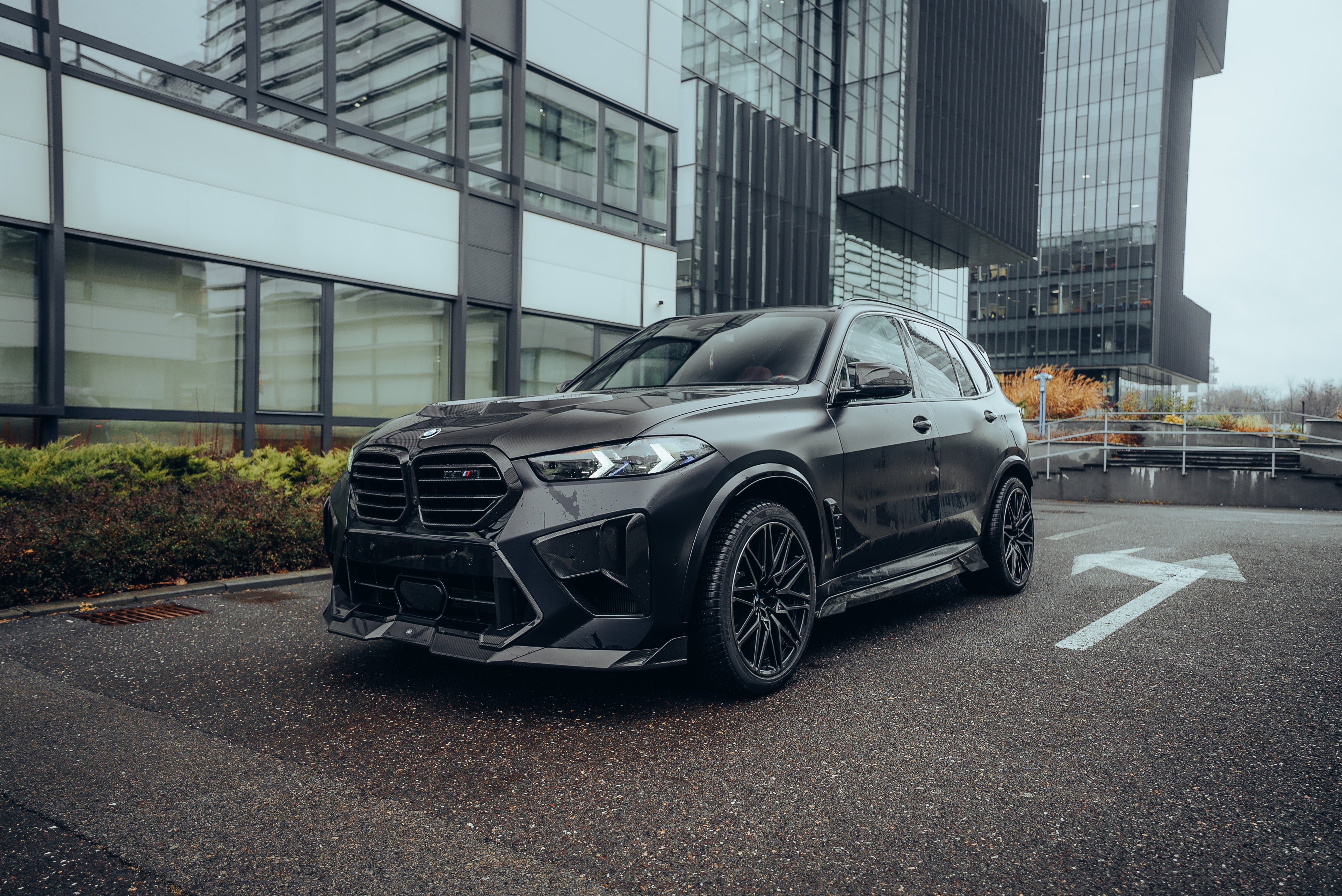 Black BMW X5M featuring a REVOZPORT carbon fiber body kit with aggressive front lip, side skirts, enlarged front air intakes, and sporty aero styling parked in a modern urban business district.
