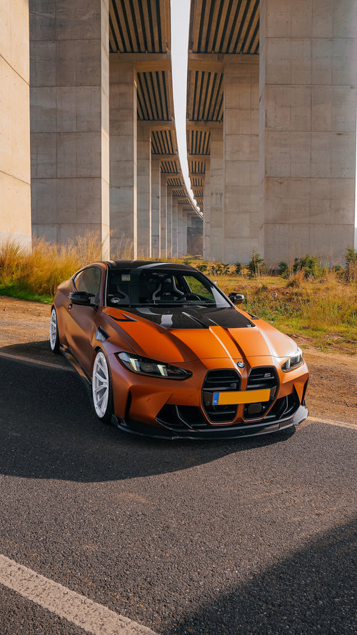 Bronze BMW G82 M4 featuring a REVOZPORT carbon fiber body kit with front lip, vented hood, side skirts, and white forged wheels parked on a scenic road near a bridge.