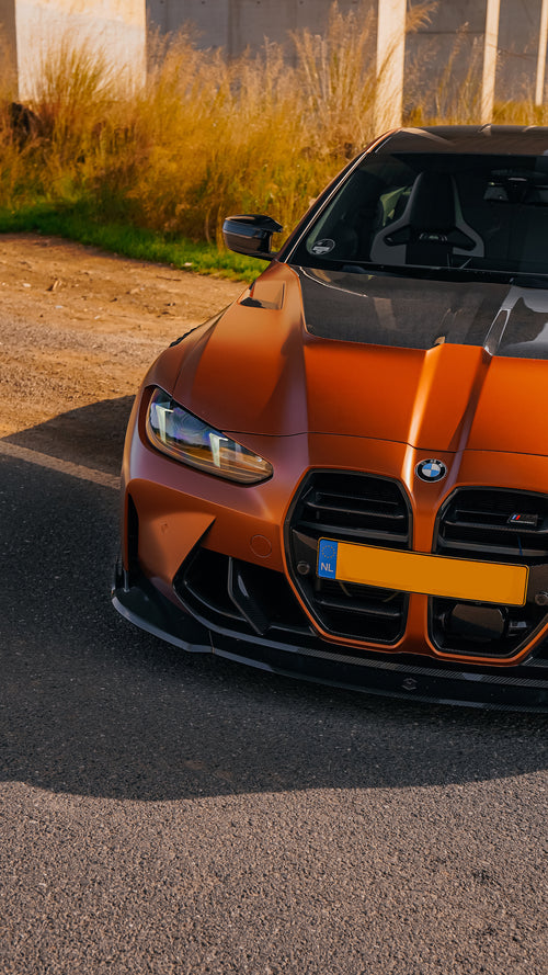 Bronze BMW G82 M4 featuring a REVOZPORT carbon fiber body kit with front lip, vented hood, side skirts, and white forged wheels parked on a scenic road near a bridge.