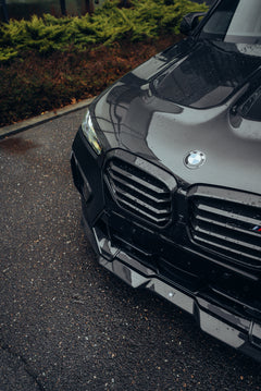 Black BMW X5M featuring a REVOZPORT carbon fiber body kit with aggressive front lip, side skirts, enlarged front air intakes, and sporty aero styling parked in a modern urban business district.