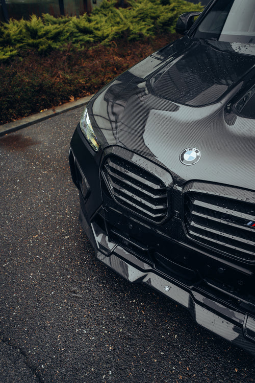 Black BMW X5M featuring a REVOZPORT carbon fiber body kit with aggressive front lip, side skirts, enlarged front air intakes, and sporty aero styling parked in a modern urban business district.