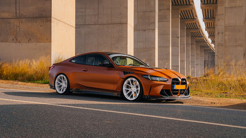 Bronze BMW G82 M4 featuring a REVOZPORT carbon fiber body kit with front lip, vented hood, side skirts, and white forged wheels parked on a scenic road near a bridge.