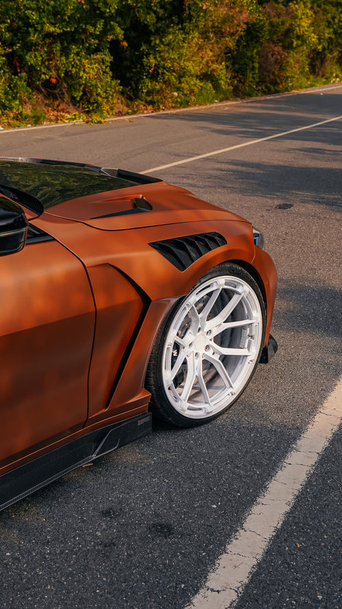 Bronze BMW G82 M4 featuring a REVOZPORT carbon fiber body kit with front lip, vented hood, side skirts, and white forged wheels parked on a scenic road near a bridge.