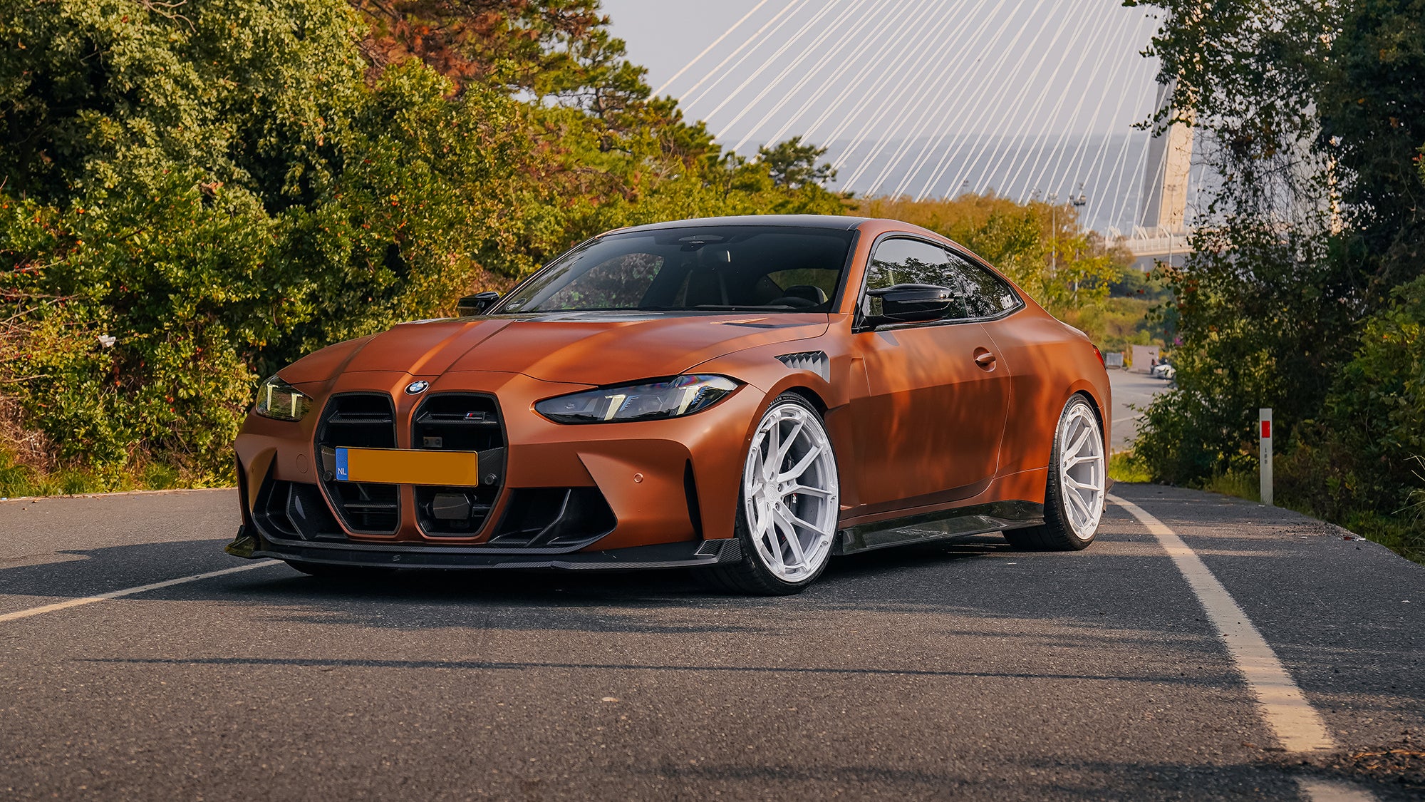 Bronze BMW G82 M4 featuring a REVOZPORT carbon fiber body kit with front lip, vented hood, side skirts, and white forged wheels parked on a scenic road near a bridge.