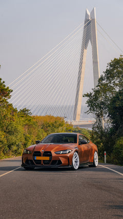 Bronze BMW G82 M4 featuring a REVOZPORT carbon fiber body kit with front lip, vented hood, side skirts, and white forged wheels parked on a scenic road near a bridge.